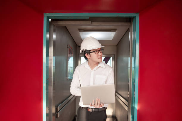 Young Asian male engineer or technician worker holding laptop computer in opened elevator door that surrounded by red wall. Mechanical engineering or building construction and maintenance concepts